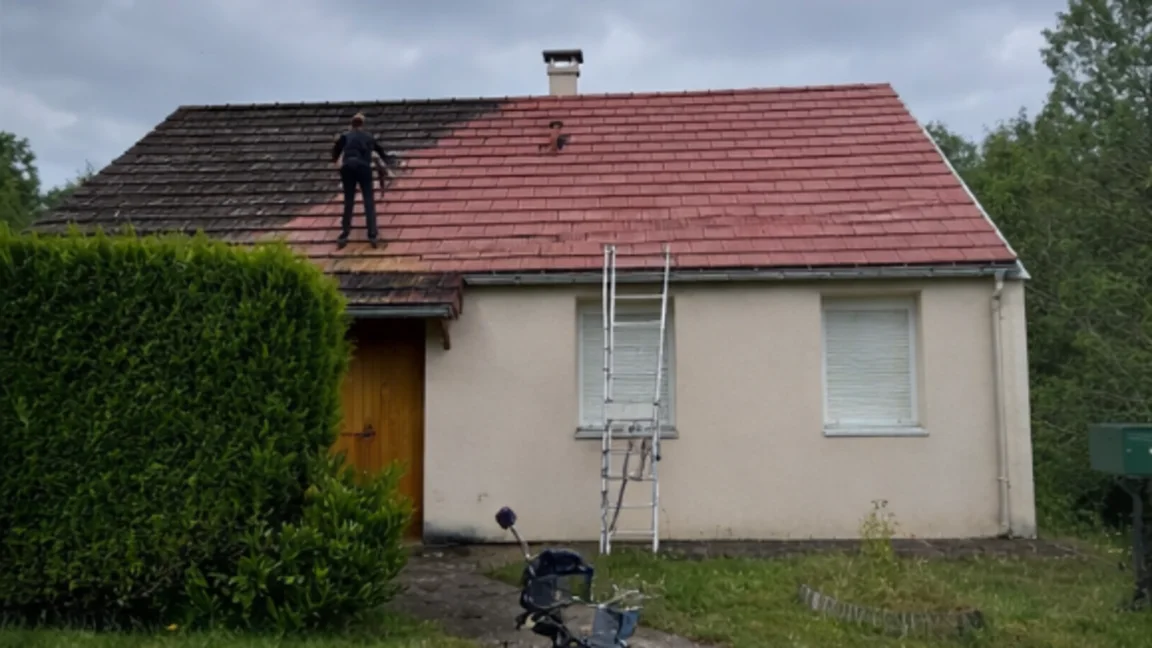 Toiture en tuiles dont la moitié est encore noircie de mousses, en cours de nettoyage.